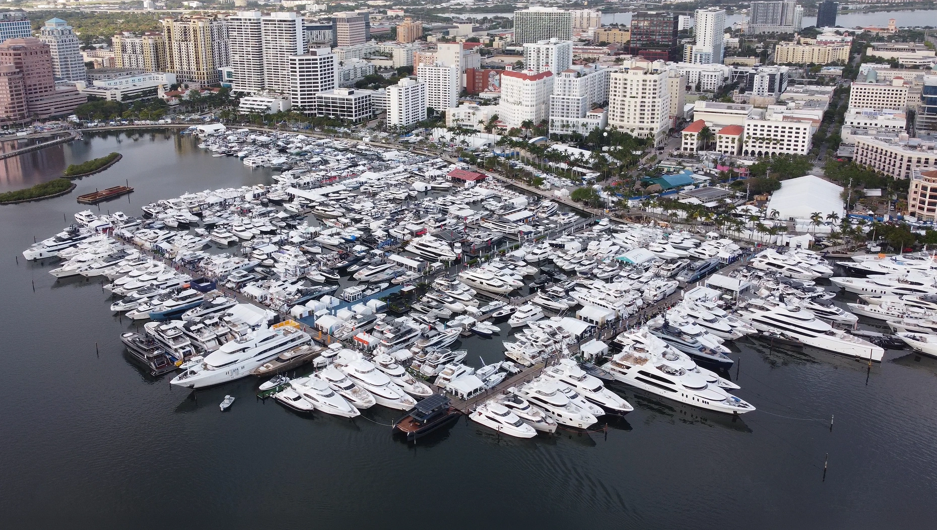 Aerial view of Palm Beach International Boat Show featuring dozens of luxury motor yachts and superyachts docked along Flagler Drive with white exhibition tents and attendees