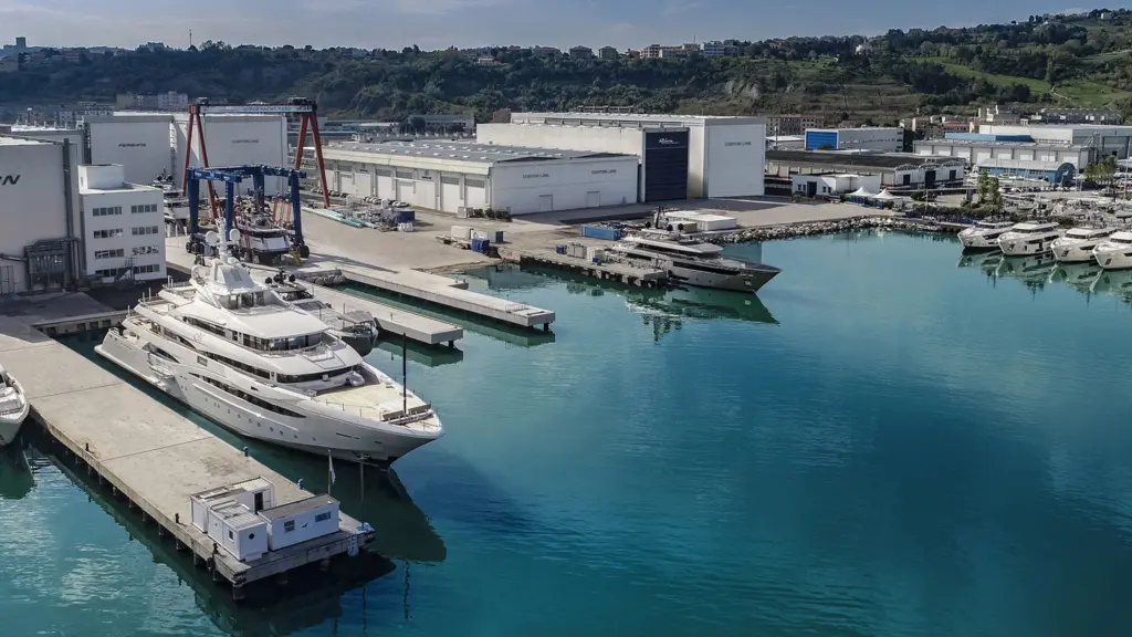 Aerial view of Riva shipyard in La Spezia, Italy showing Custom Line production facilities, superyachts at dock including white hull vessels, and turquoise harbor waters