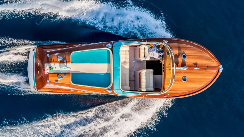 Aerial view of classic Riva Aquarama wooden speedboat with iconic mahogany deck, turquoise upholstery, and white leather seating cutting through deep blue water with wake